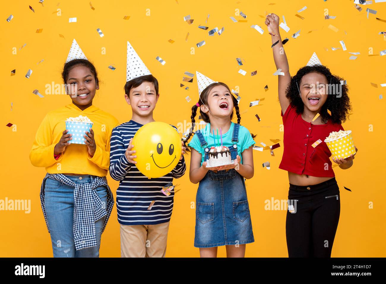 Multiethnische Kinder feiern Geburtstagsfeier im gelben Farbenhintergrund des Studios mit Konfetti Stockfoto