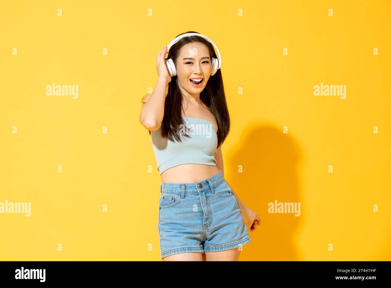 Junge hübsche asiatische Frau, die Kopfhörer trägt, Musik hört und tanzt im gelben Hintergrund des Studios Stockfoto