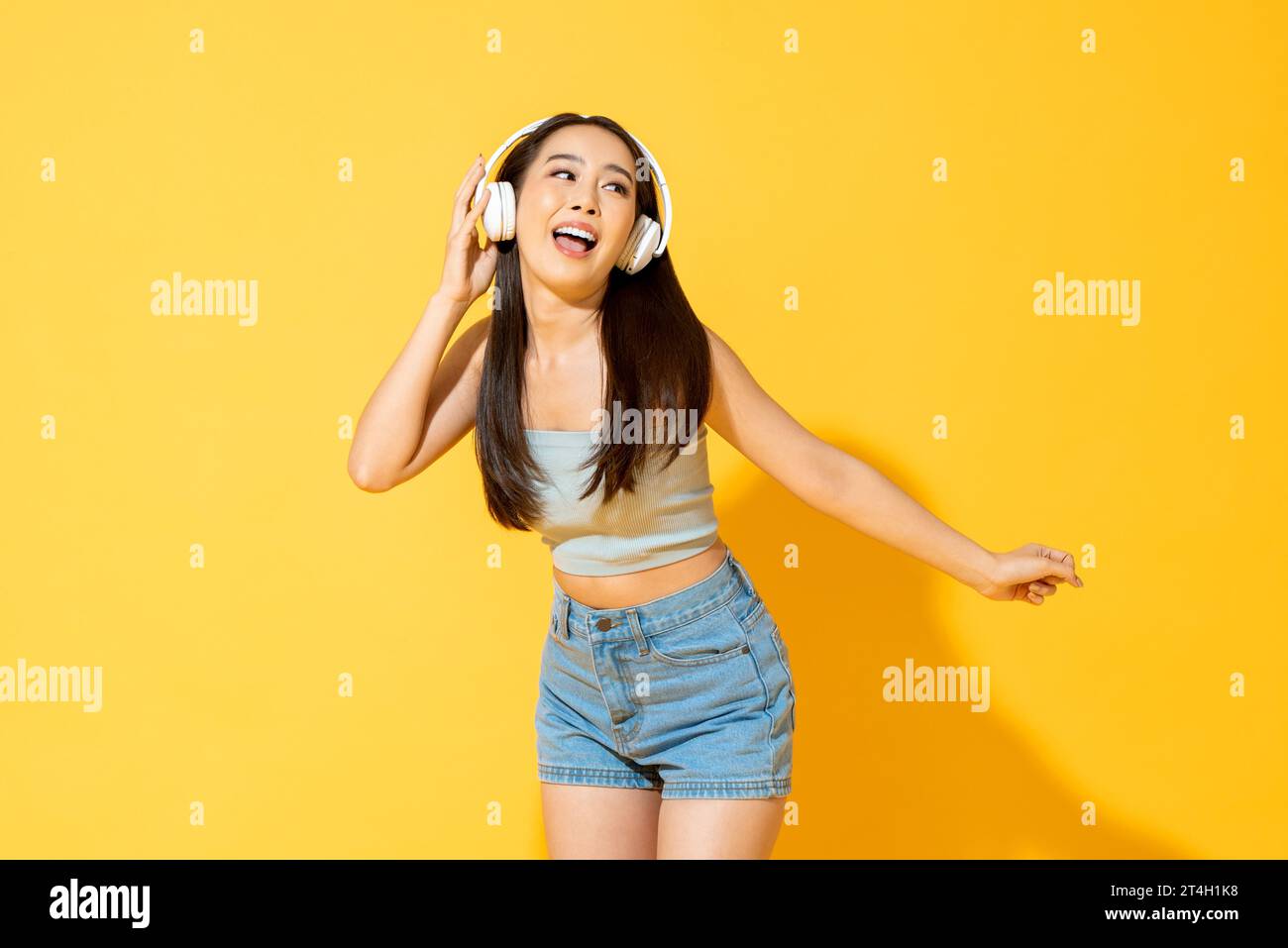 Junge hübsche asiatische Frau, die Kopfhörer trägt, Musik hört und tanzt im gelben Hintergrund des Studios Stockfoto