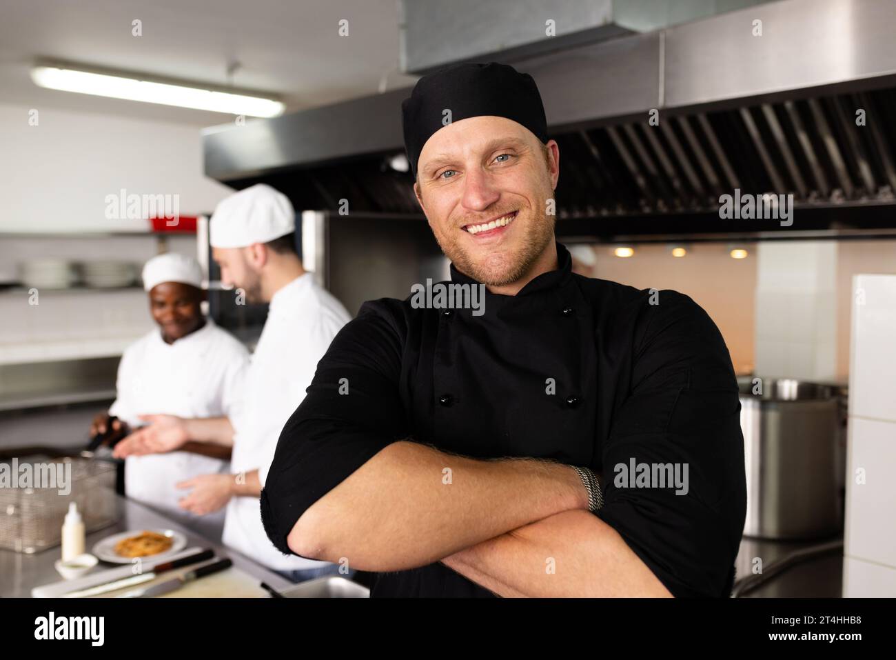 Vielseitig lächelnder männlicher Chefkoch mit überkreuzten Armen, der vor Studenten steht, die im Hintergrund kochen Stockfoto