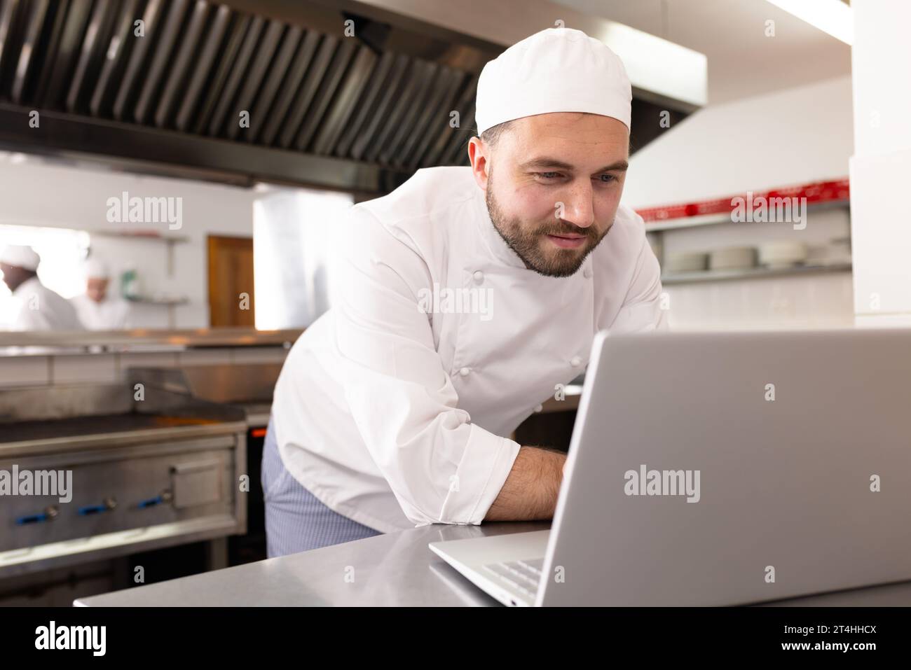 Kaukasischer Küchenchef in weißer Uniform mit Laptop auf der Theke in der Geschäftsküche Stockfoto