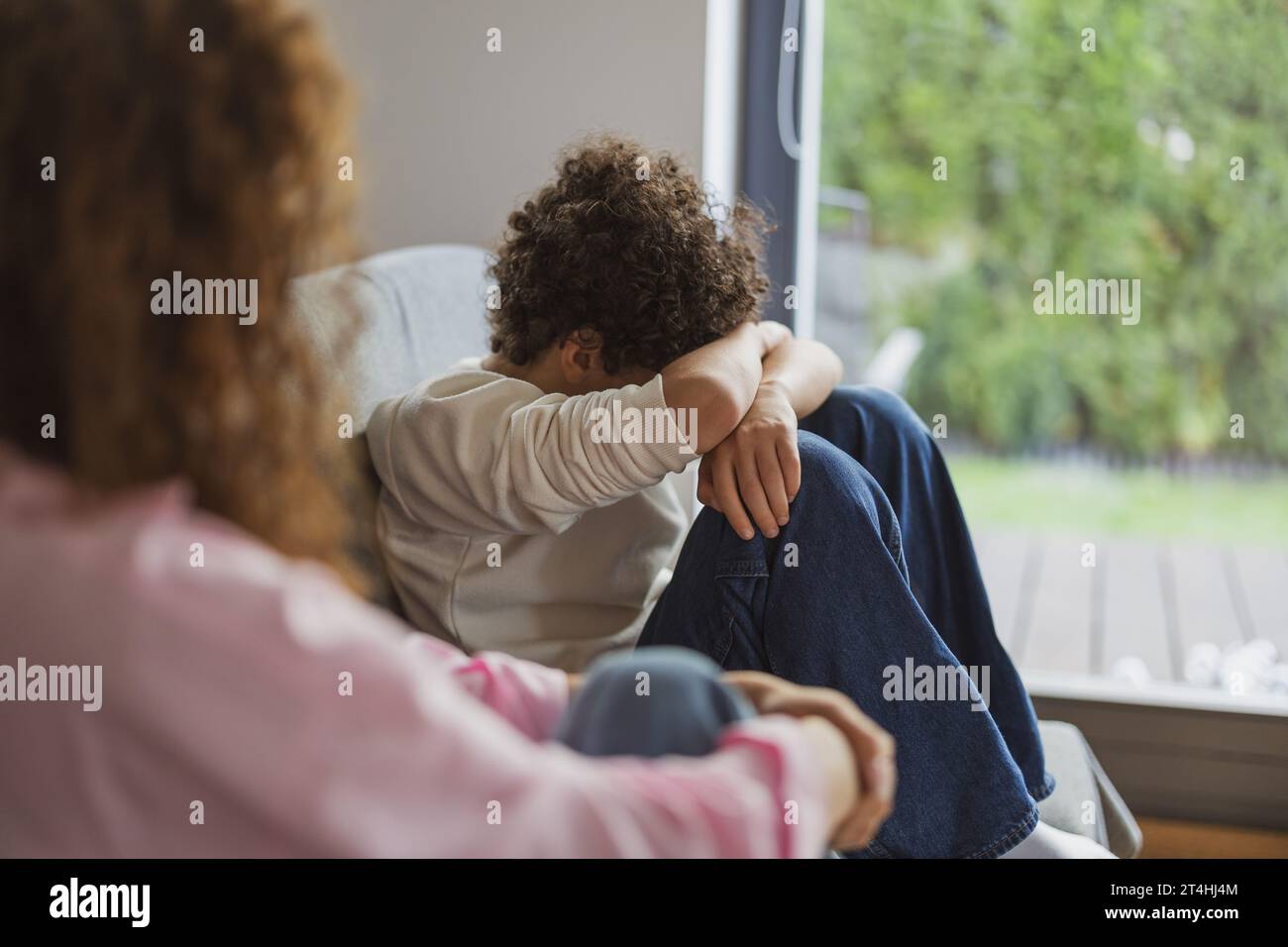 Ein verärgerter Junge sitzt auf dem Sofa, während seine Mutter ihn ansieht Stockfoto