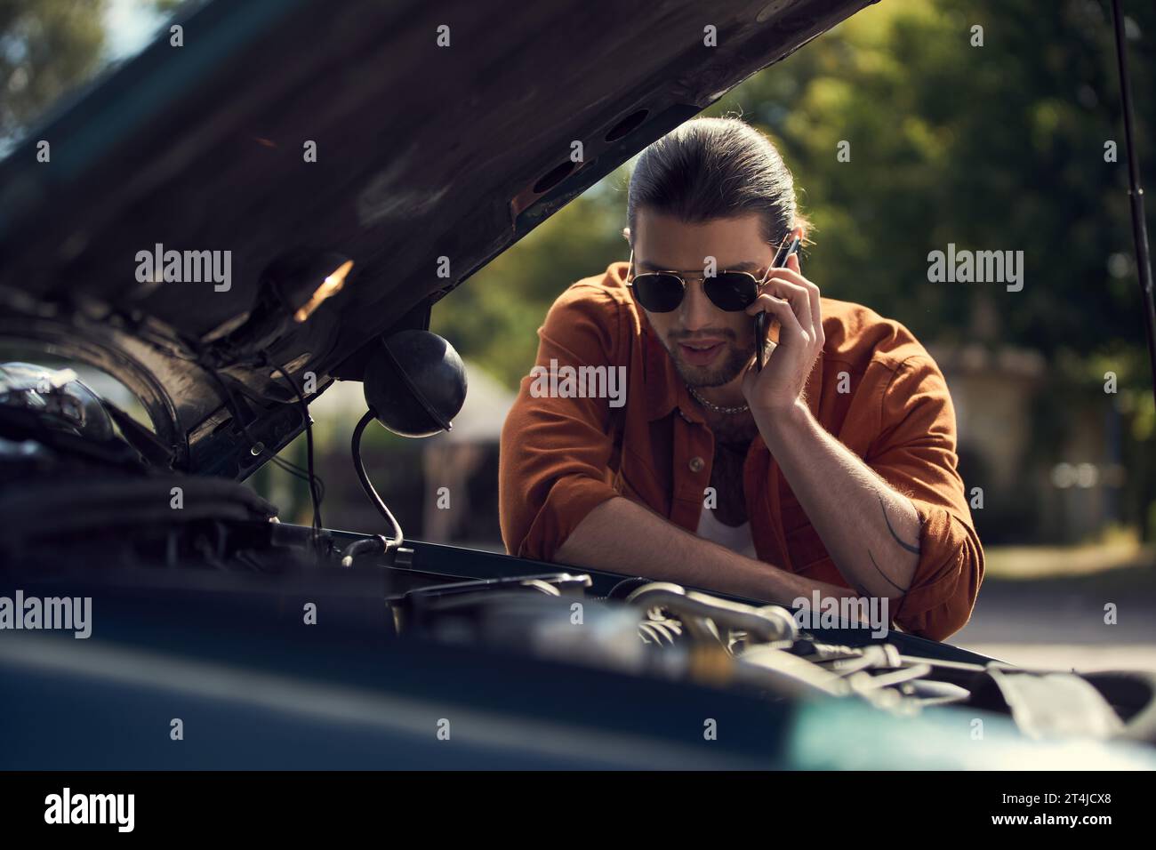 Ein stilvoller Fahrer in eleganter Kleidung ruft seinen Versicherer an und blickt auf die geöffnete Motorhaube seines Autos Stockfoto