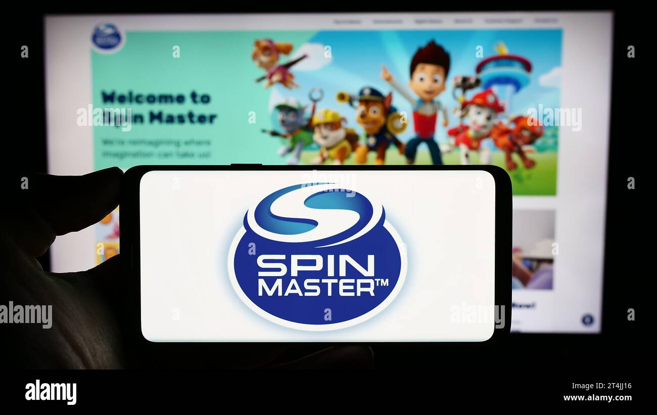 Person mit Handy mit Logo des kanadischen Spielzeugunternehmens Spin Master Corp. Vor der Webseite. Schwerpunkt auf dem Display des Telefons. Stockfoto