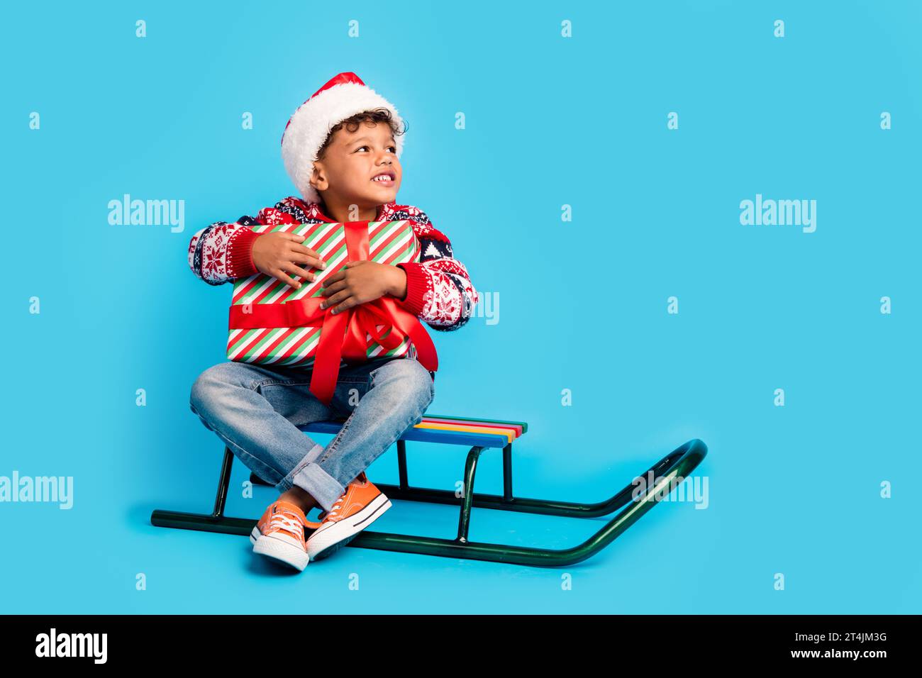 Foto in voller Länge von Dreamy Boy gekleidet Pullover santa Hut sitzen auf Schlitten Umarmung Geschenk Blick leer Raum isoliert auf blauem Hintergrund Stockfoto