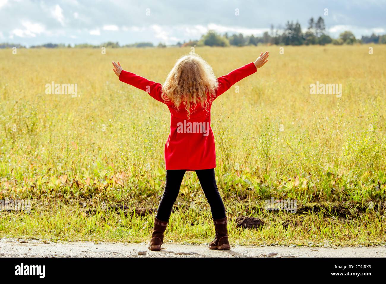 Rückansicht eines 8-jährigen Mädchens mit rotem Mantel, lockigen blonden Haaren, stehend auf einer Wiese, wunderschöner Blick auf die Natur im Freien. Stockfoto