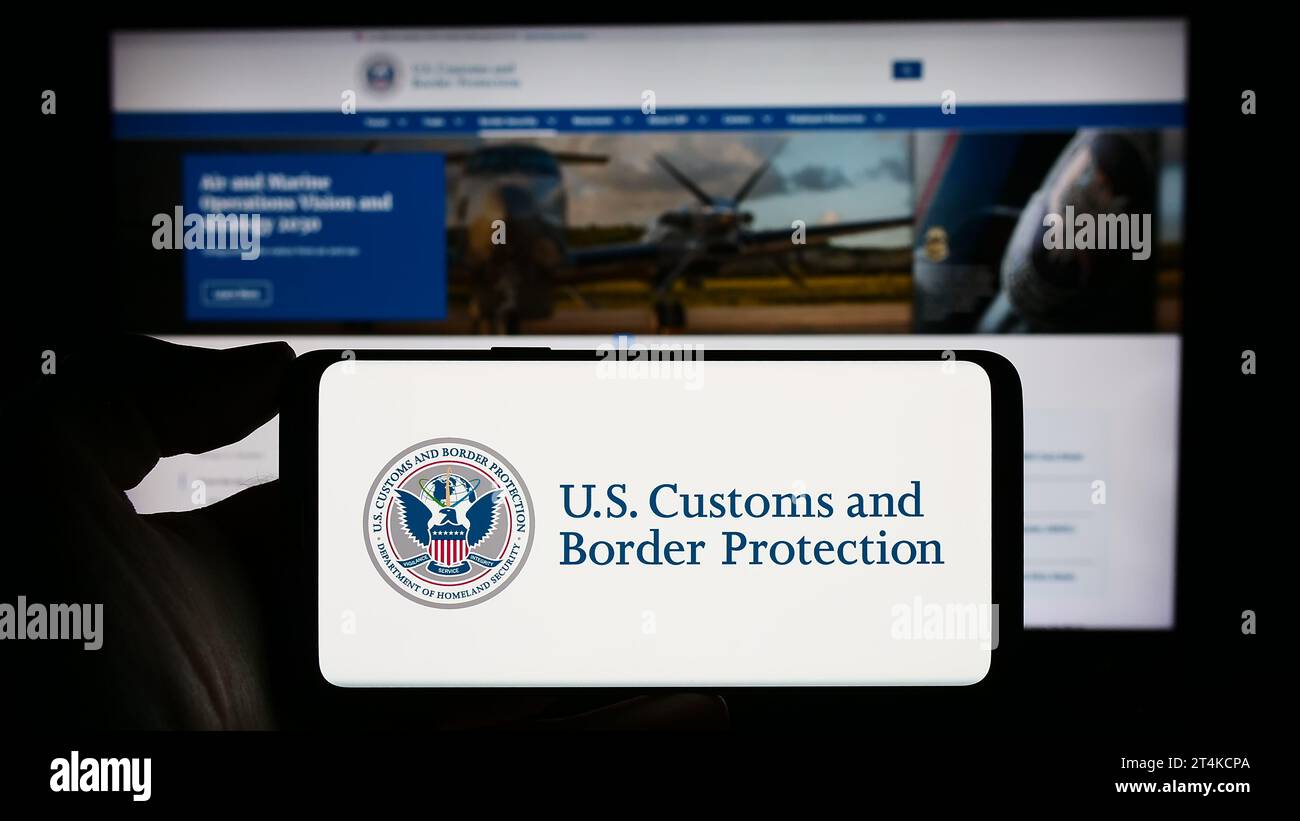 Person, die ein Mobiltelefon mit dem Zollsiegel der Vereinigten Staaten (United States Customs and Border Protection, CBP) vor der Webseite hält. Schwerpunkt auf dem Display des Telefons. Stockfoto