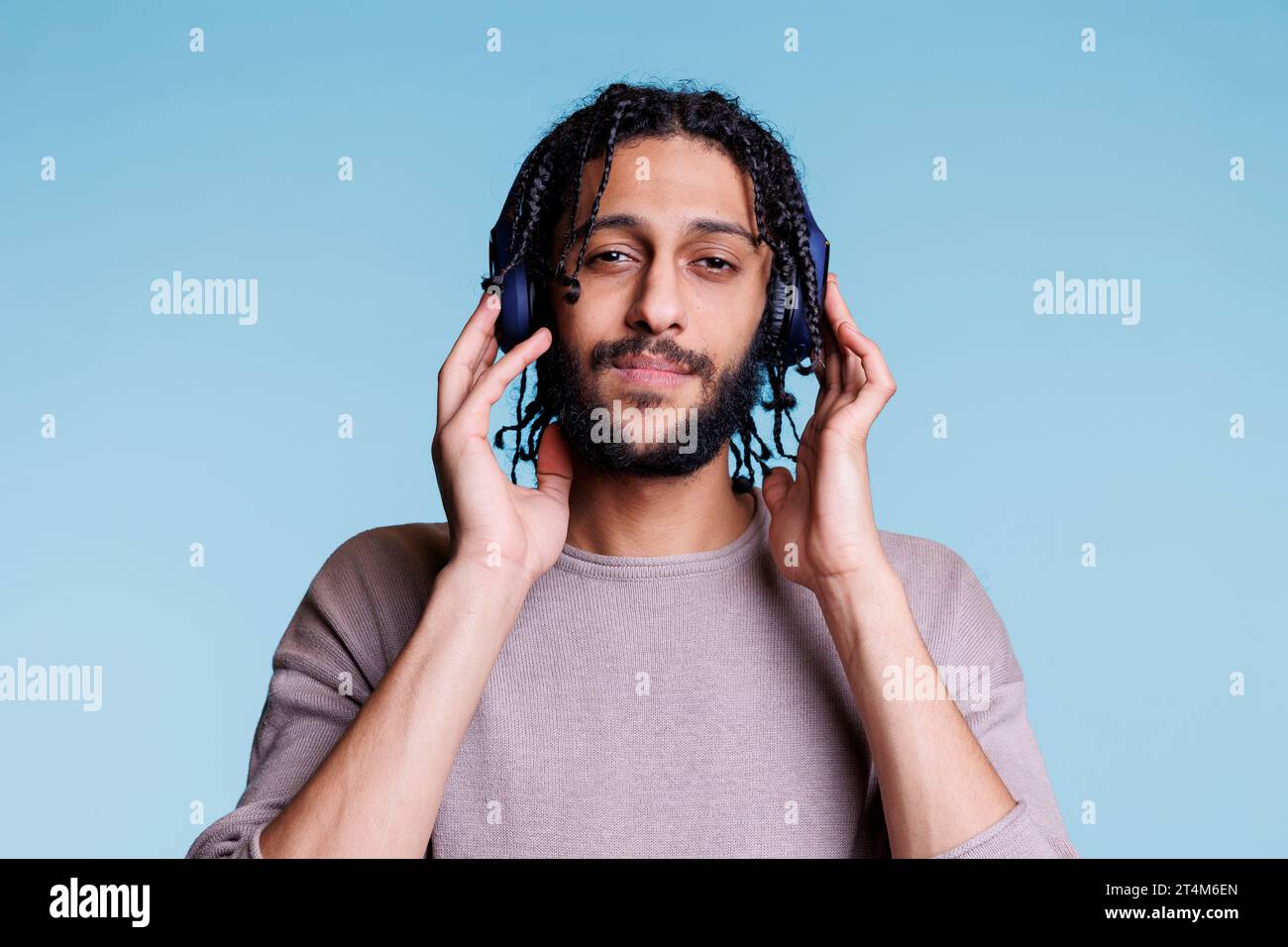 Junger arabischer Mann, der Kopfhörer auf dem Kopf berührt, während er Musik hört, lächelt und in die Kamera blickt. Person, die schnurlose Ohrhörer trägt, Sound genießt und Porträt entspannt Stockfoto