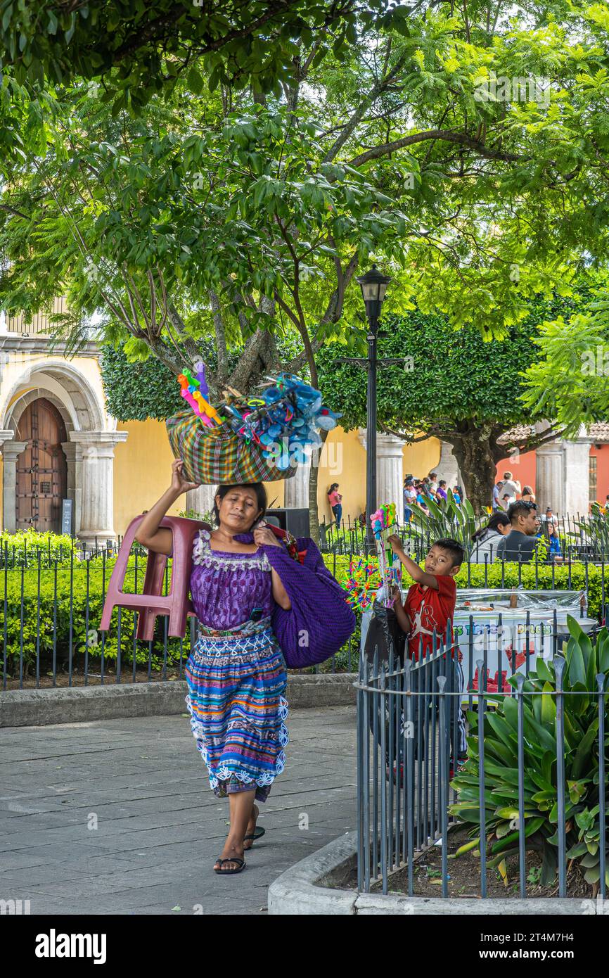 Guatemala, La Antigua - 20. Juli 2023: Famale-Verkäuferin in farbenfroher traditioneller Kleidung spaziert auf dem Plaza Mayor, dem zentralen Platz, mit einem Korb mit Spielzeug auf dem Kopf Stockfoto
