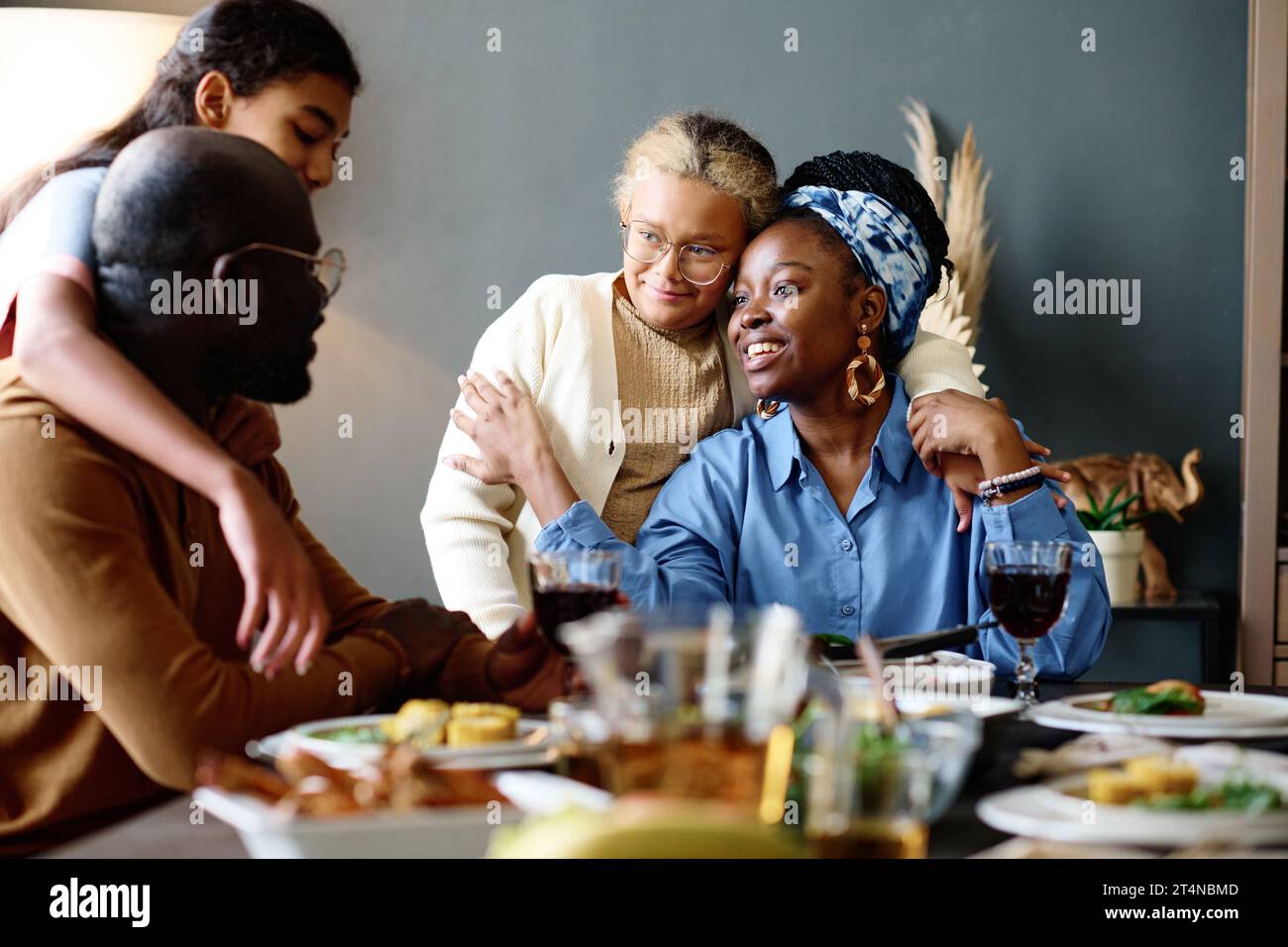 Zwei liebevolle Töchter, die ihren Vater und ihre Mutter umarmen, sitzen am festlichen Tisch, serviert mit hausgemachten Speisen und Getränken zum Abendessen Stockfoto
