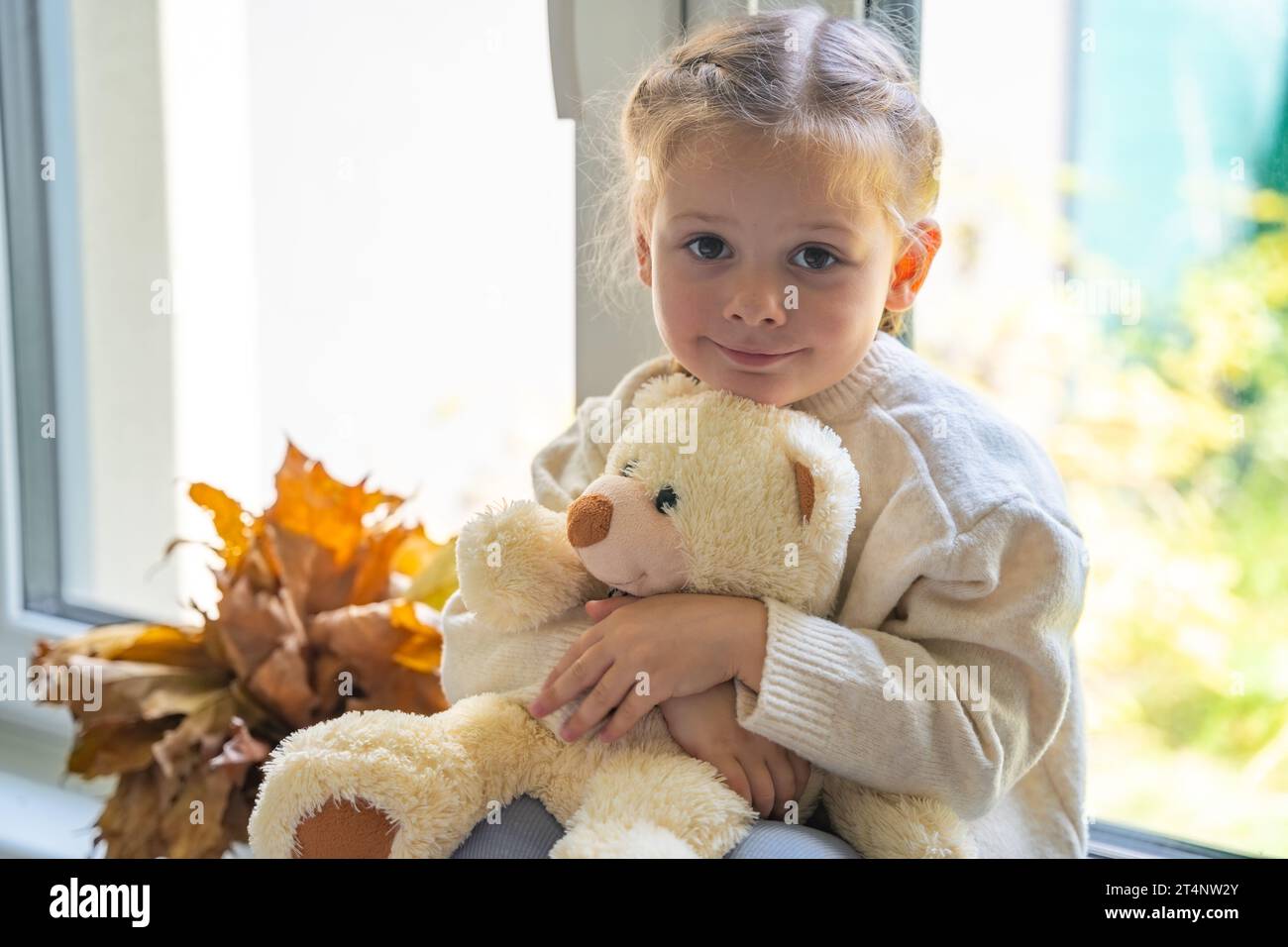 Porträt eines kleinen Mädchens im warmen Pullover, das im Herbst mit dem kleinen Teddybären am Fenster sitzt Stockfoto