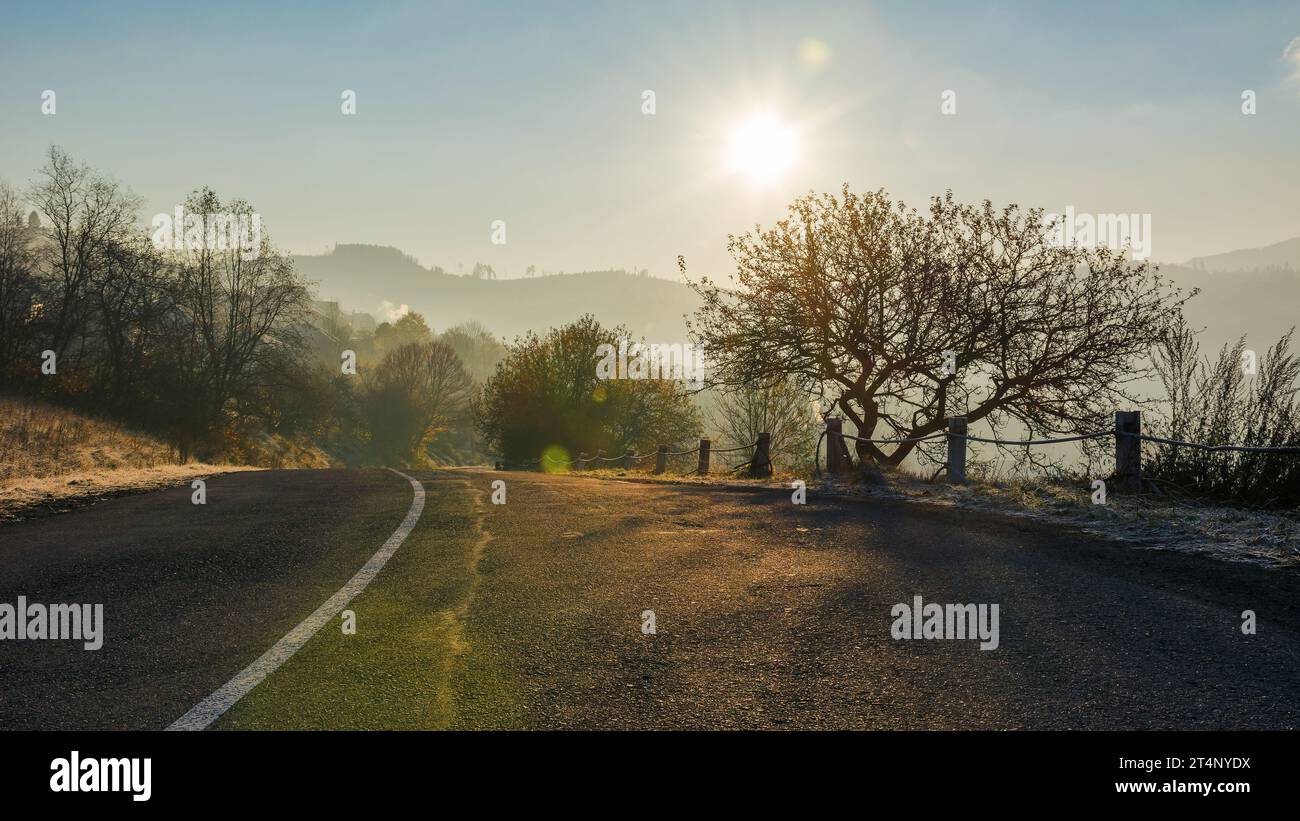 Straße durch bergige Landschaft im Morgenlicht. Blattloser Baum auf den Hügeln. Sonne am blauen wolkenlosen Himmel Stockfoto