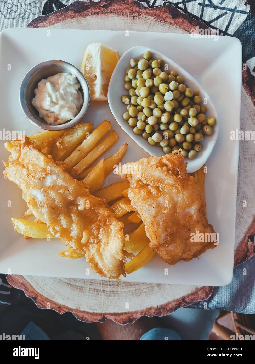 Vertikales Foto von Draufsicht von Fisch mit Pommes Frites und grünen Erbsen auf einer weißen quadratischen Platte mit Saucen. Weichzeichner. Kopierbereich Stockfoto