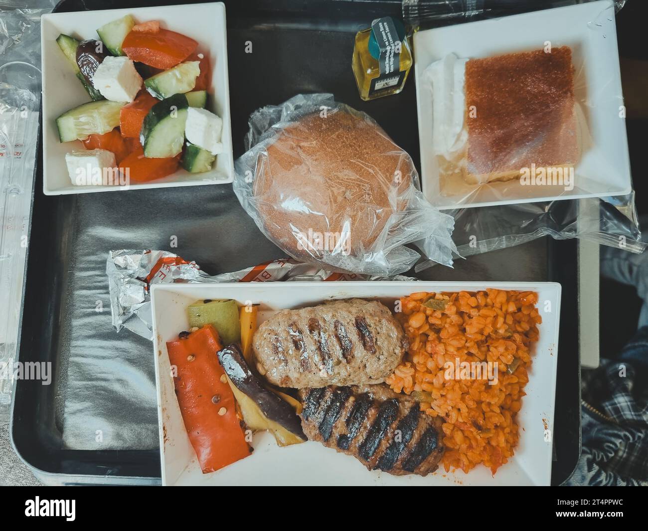 Draufsicht auf ein Tablett mit Lebensmitteln in einem Flugzeug. Zwei Schnitzel mit Bulgur, ein Brötchen, Olivenöl und frischem Gemüsesalat. Weichzeichner. Stockfoto