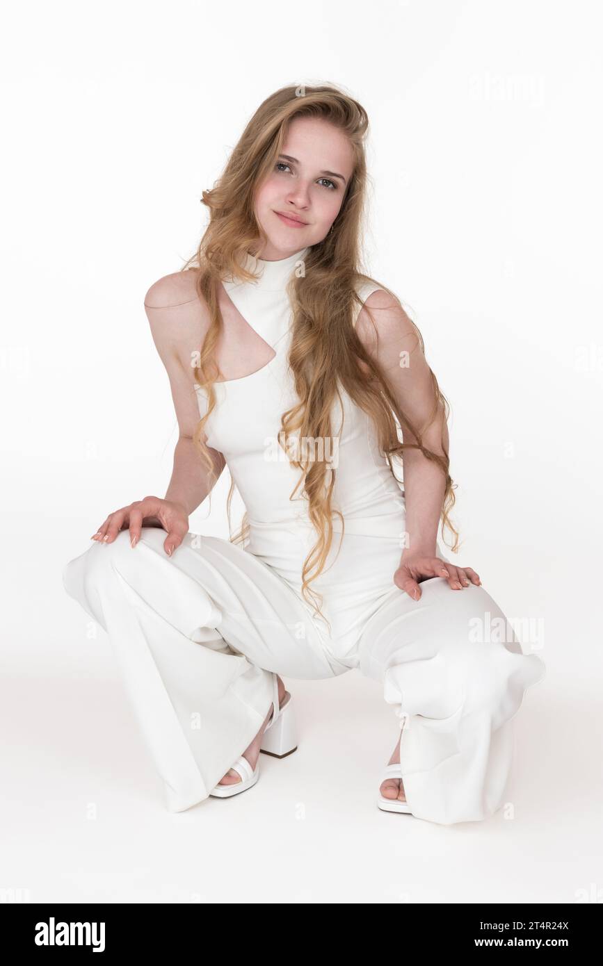 Vorderansicht einer jungen blonden Frau in weißem Jumpsuit und Slingbacks, die in der Hockposition sitzt, beide Hände auf die Knie legte und in die Kamera schaute Stockfoto
