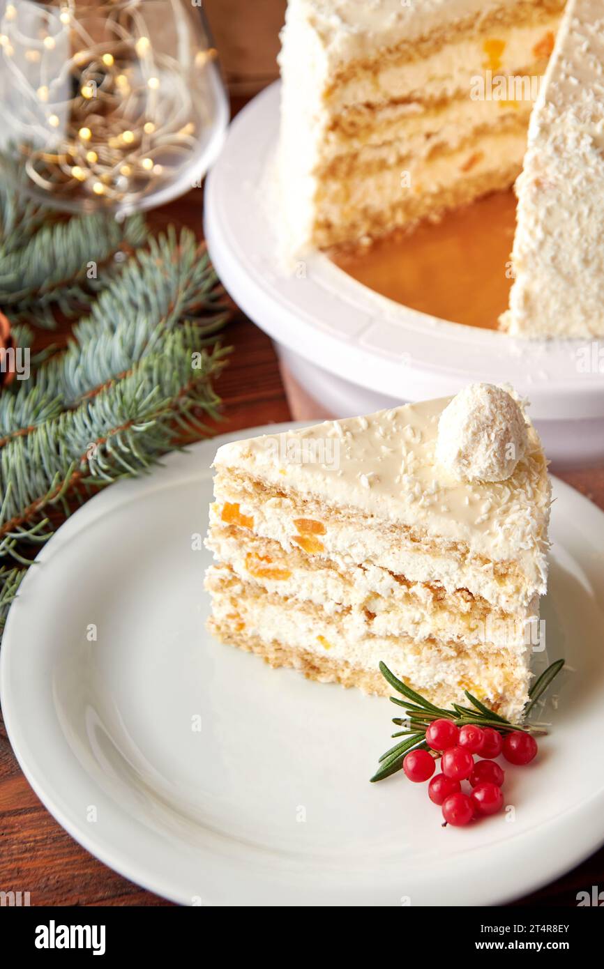 Ein Stück festlicher Weihnachtskuchen mit Kokos und Pfirsichen, dekoriert mit Preiselbeeren und Rosmarin auf weißem Teller. Stockfoto