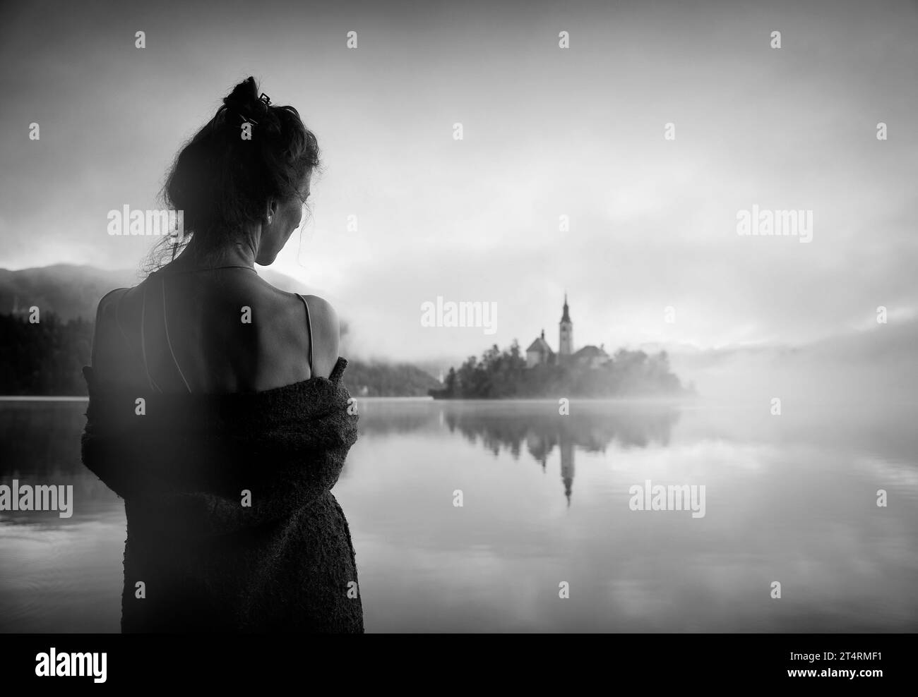 Frau im grauen Mantel, die einen Sonnenaufgang im Nebel am Ufer des Bleder Sees beobachtet, Slowenien II Stockfoto