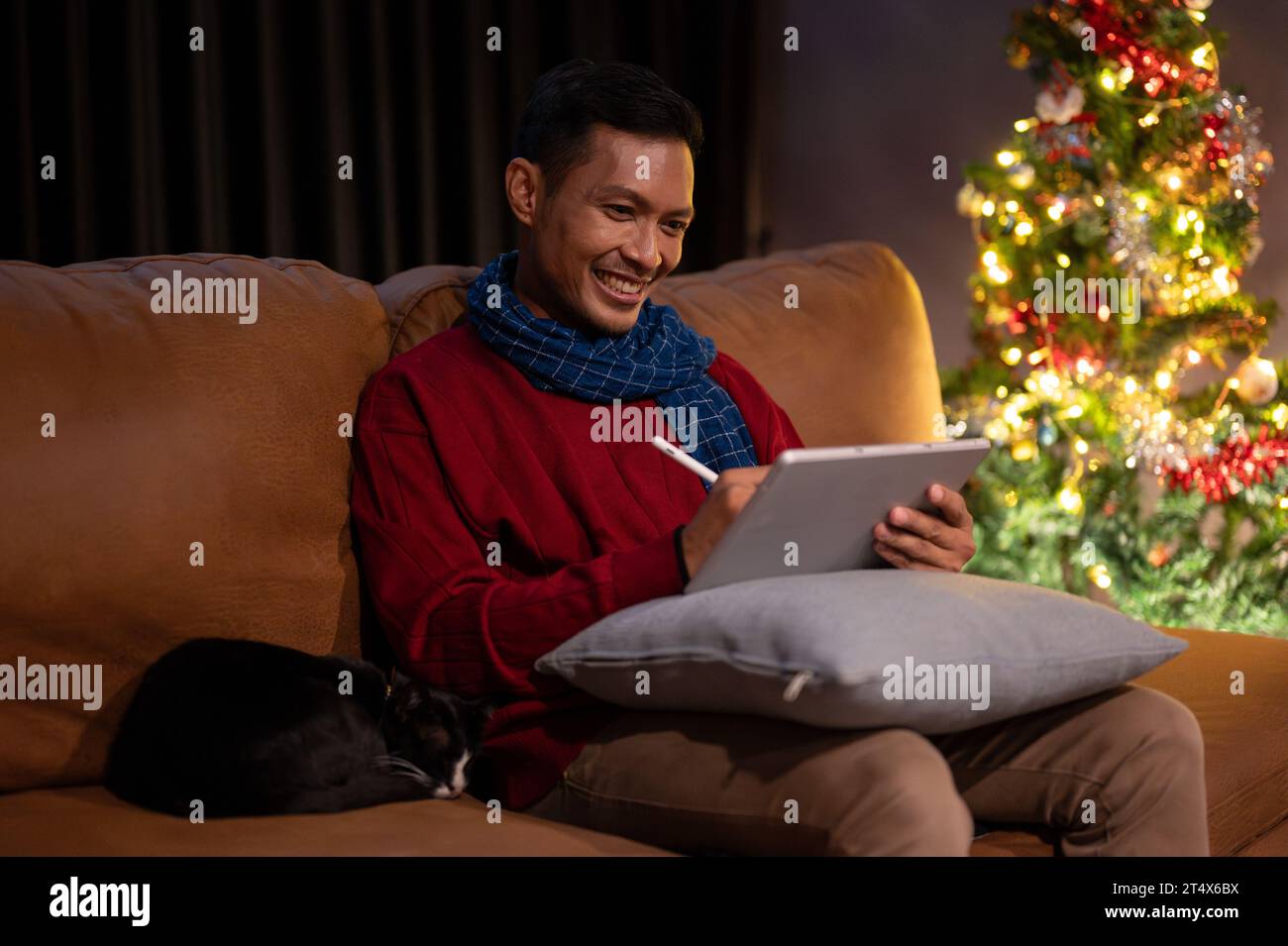 Ein glücklicher asiatischer Mann benutzt sein digitales Tablet auf einer Couch, während seine liebenswerte Katze neben ihm schläft und zusammen Weihnachtsnacht zu Hause verbringt. Stockfoto