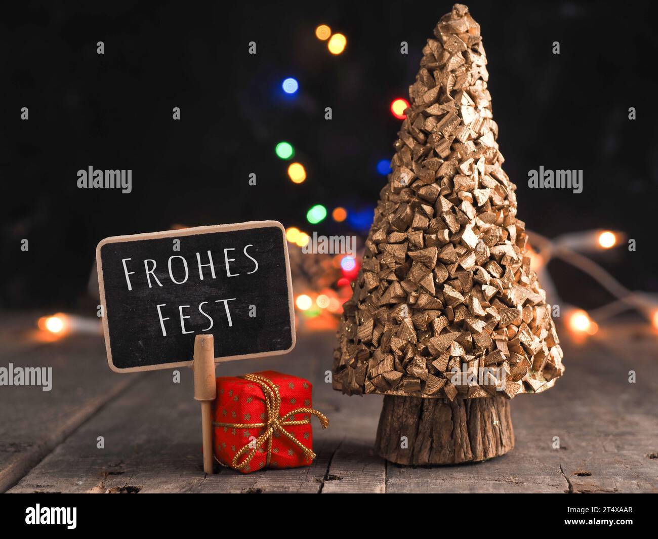 Kleine Tafel mit der deutschen Aufschrift Frohe Weihnachten auf einem Holztisch, verschwommene Weihnachtslichter im Hintergrund Stockfoto