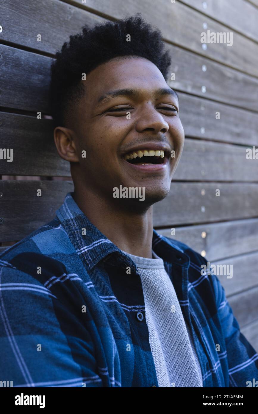 Porträt eines glücklichen afroamerikanischen Mannes in blauem kariertem Hemd, der gegen Holzzaun lacht Stockfoto