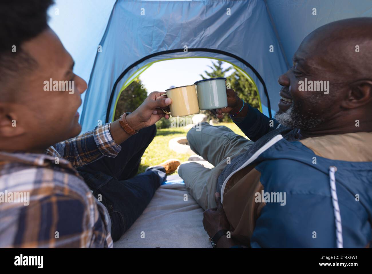 Glücklicher afroamerikanischer Vater und erwachsener Sohn im Zelt trinken einen Toast mit Tassen Kaffee Stockfoto