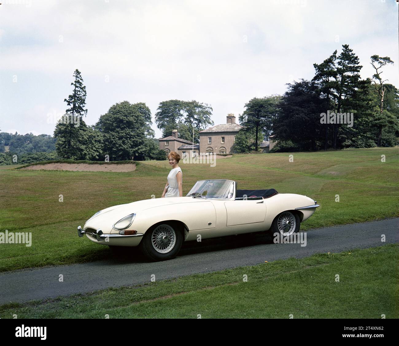Frühe Werbefoto des Jaguar E-TYPE S1, offener Zweisitzer, 3,8 L, mit Modell in weißem Kleid im Halesowen Golf Club mit Clubhaus im Hintergrund. Stockfoto
