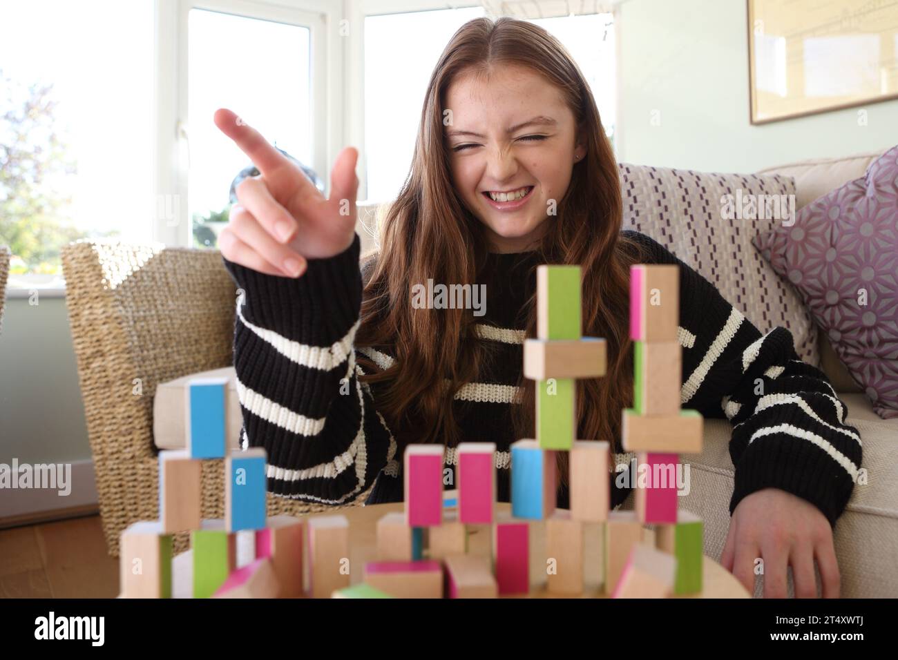 Ein Mädchen im Teenageralter lächelt und lacht, zerstört eine Mauer und Türme aus Holzblöcken und schlägt das Muster mit der Hand nieder Stockfoto