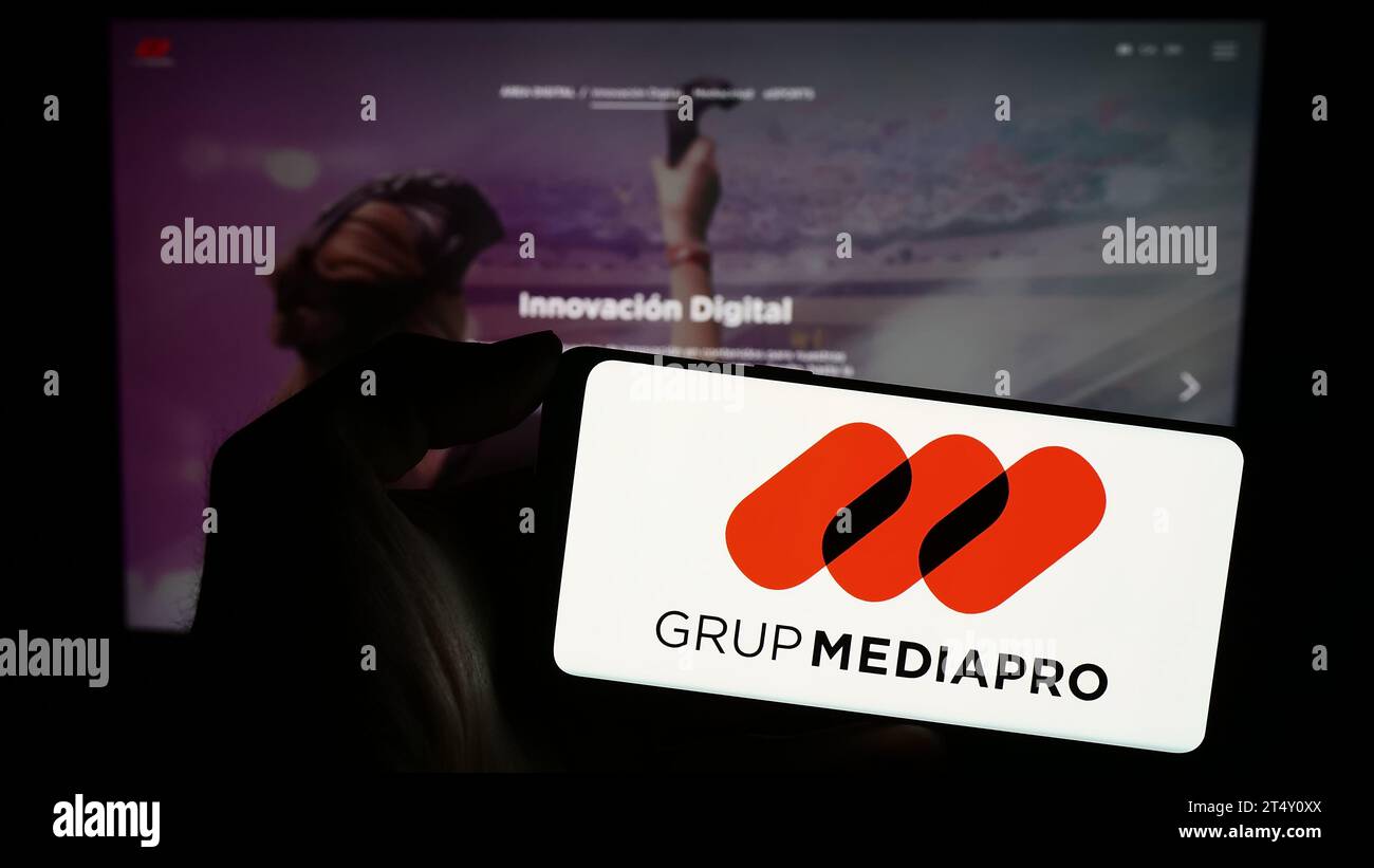 Person, die ein Mobiltelefon mit dem Logo des spanischen Multimedia-Unternehmens Mediapro Group vor der Business-Webseite hält. Schwerpunkt auf dem Display des Telefons. Stockfoto