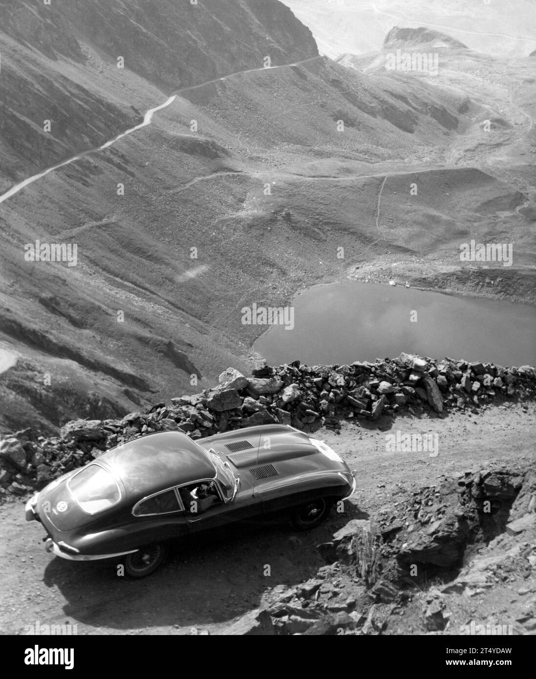 Pressefoto von 1961 E-TYPE, S1, Fixed Head Coupe, 6162RW auf einem Schotterweg Bergpass mit See und Bergstraße im Hintergrund. Stockfoto