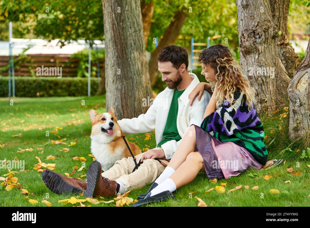 Glücklicher Mann und lockige Frau in süßem Outfit, die Corgi-Hund anschauen und in der Nähe eines Baumes im herbstlichen Park sitzen Stockfoto