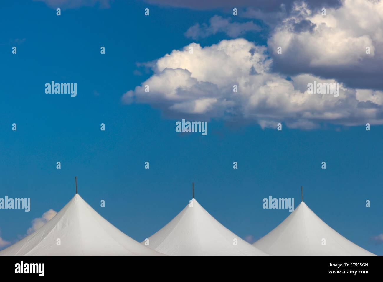Weiße flauschige Wolken im hellblauen Himmel ziehen über die Spitzen von drei weißen Zelten. Stockfoto