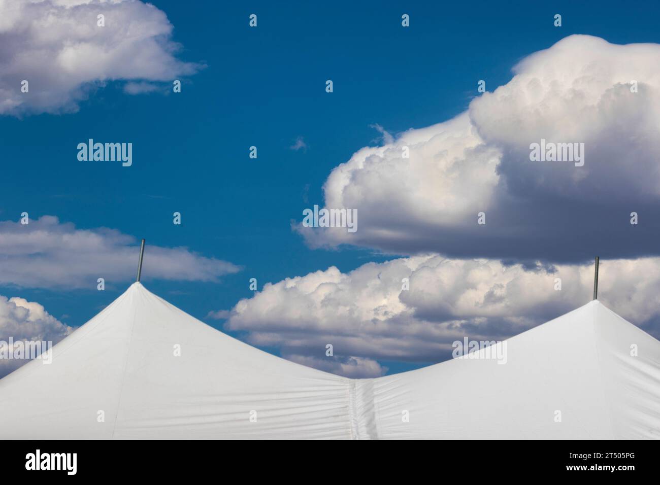 Weiße flauschige Wolken in hellblauem Himmel hoover über den Spitzen eines weißen Zeltes. Stockfoto