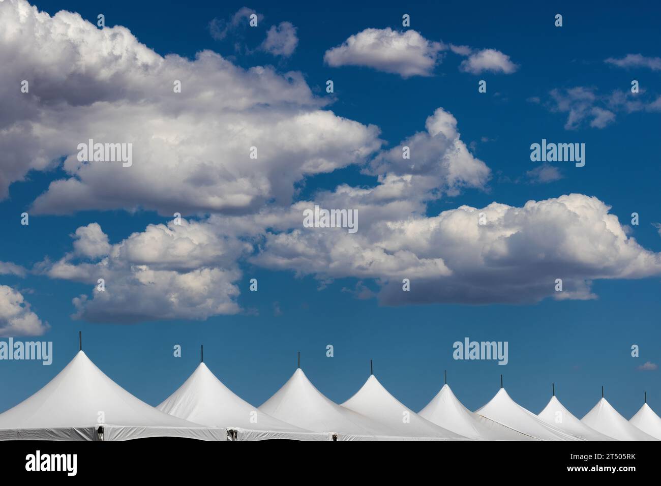 Große weiße flauschige Wolken am blauen Himmel über einer Linie weißer Zeltspitzen. Stockfoto
