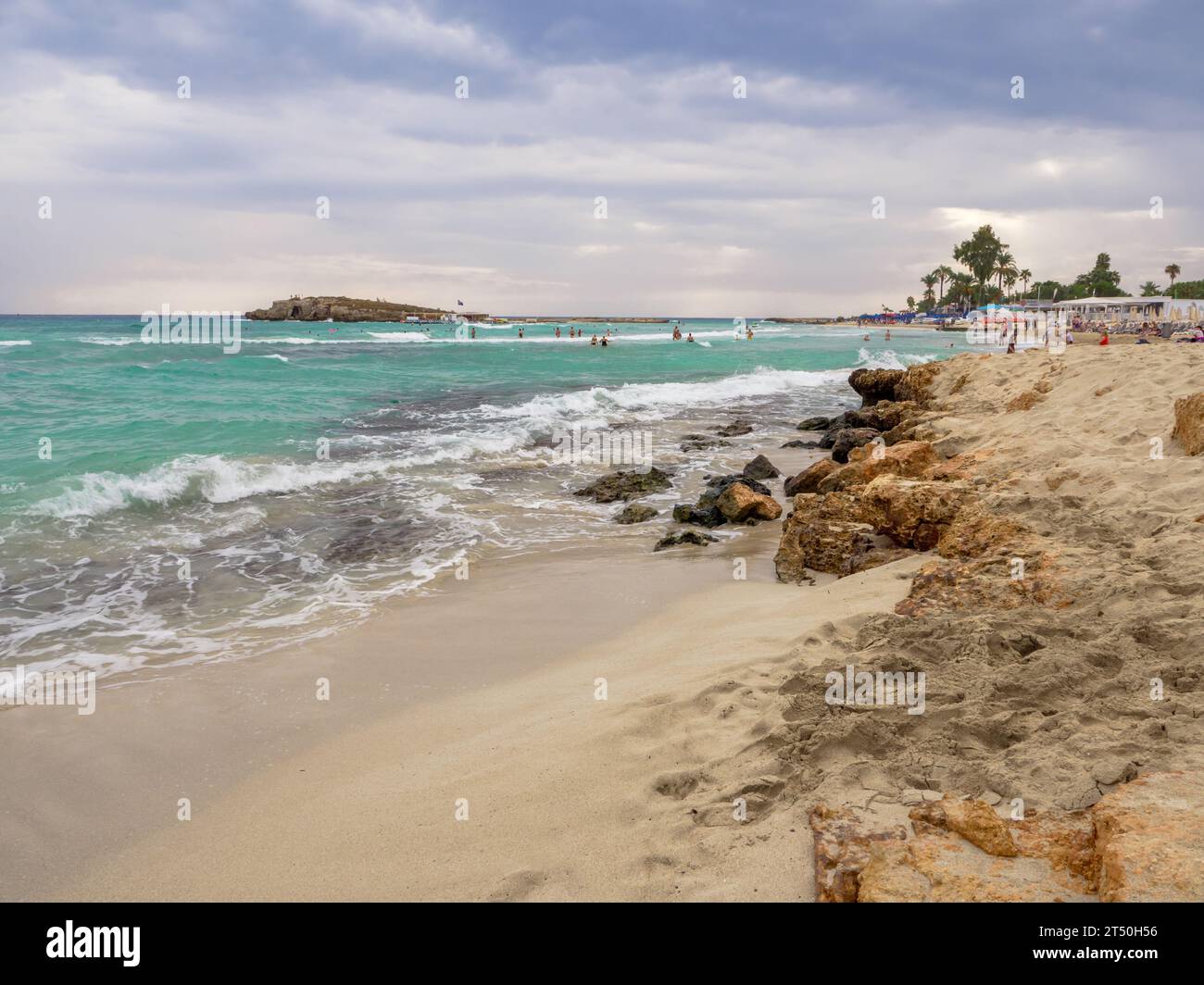 Menschen, die an bewölkten Tagen in Meereswellen schwimmen. Malerischer Nissi-Strand mit turbulentem Cyan-Wasser, eine kleine felsige Insel, bewölkter Himmel in Ayia Napa, Zypern Stockfoto