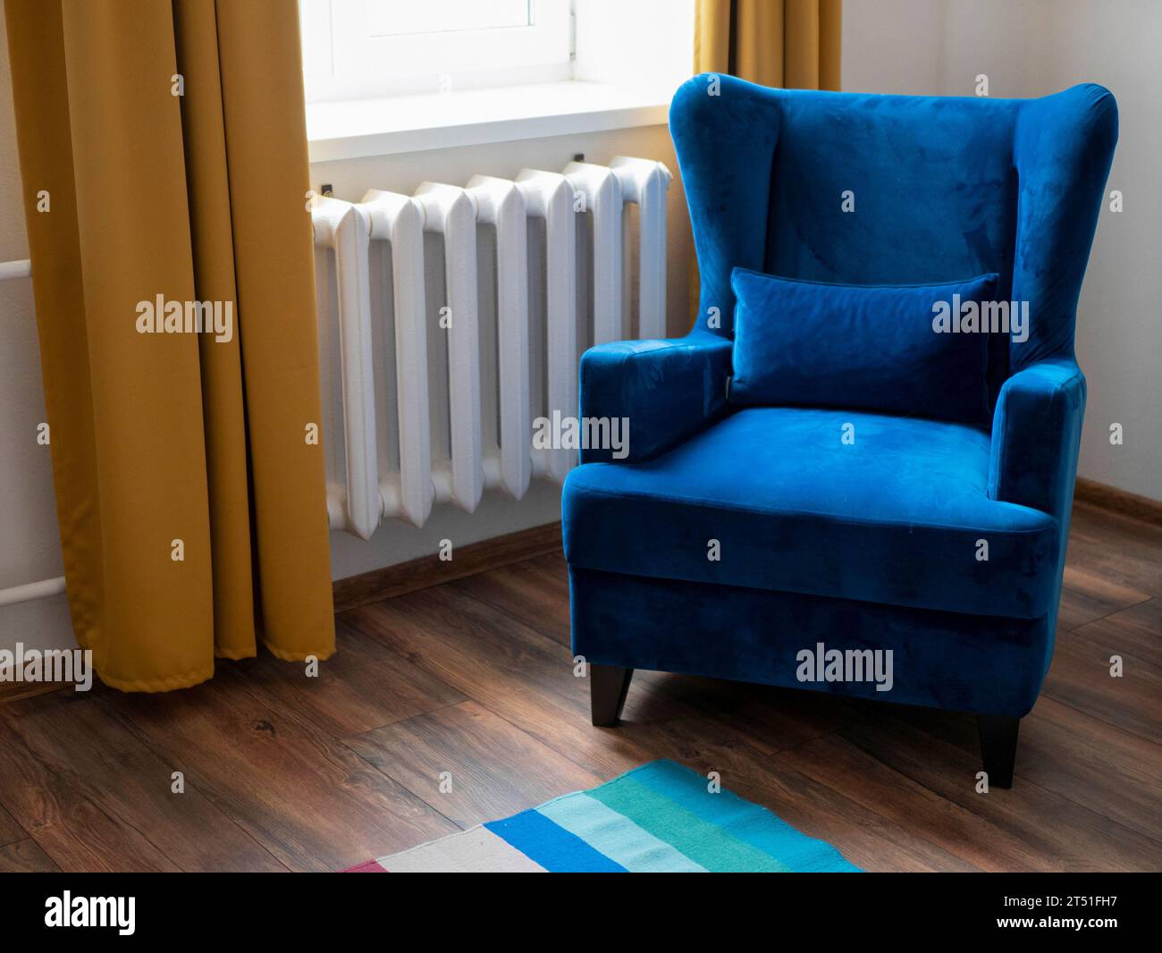 Wohnzimmerteil mit blauem Sessel, Fenster und gelbem Vorhang, Holzboden und gestreiftem Teppich, selektiver Fokus Stockfoto