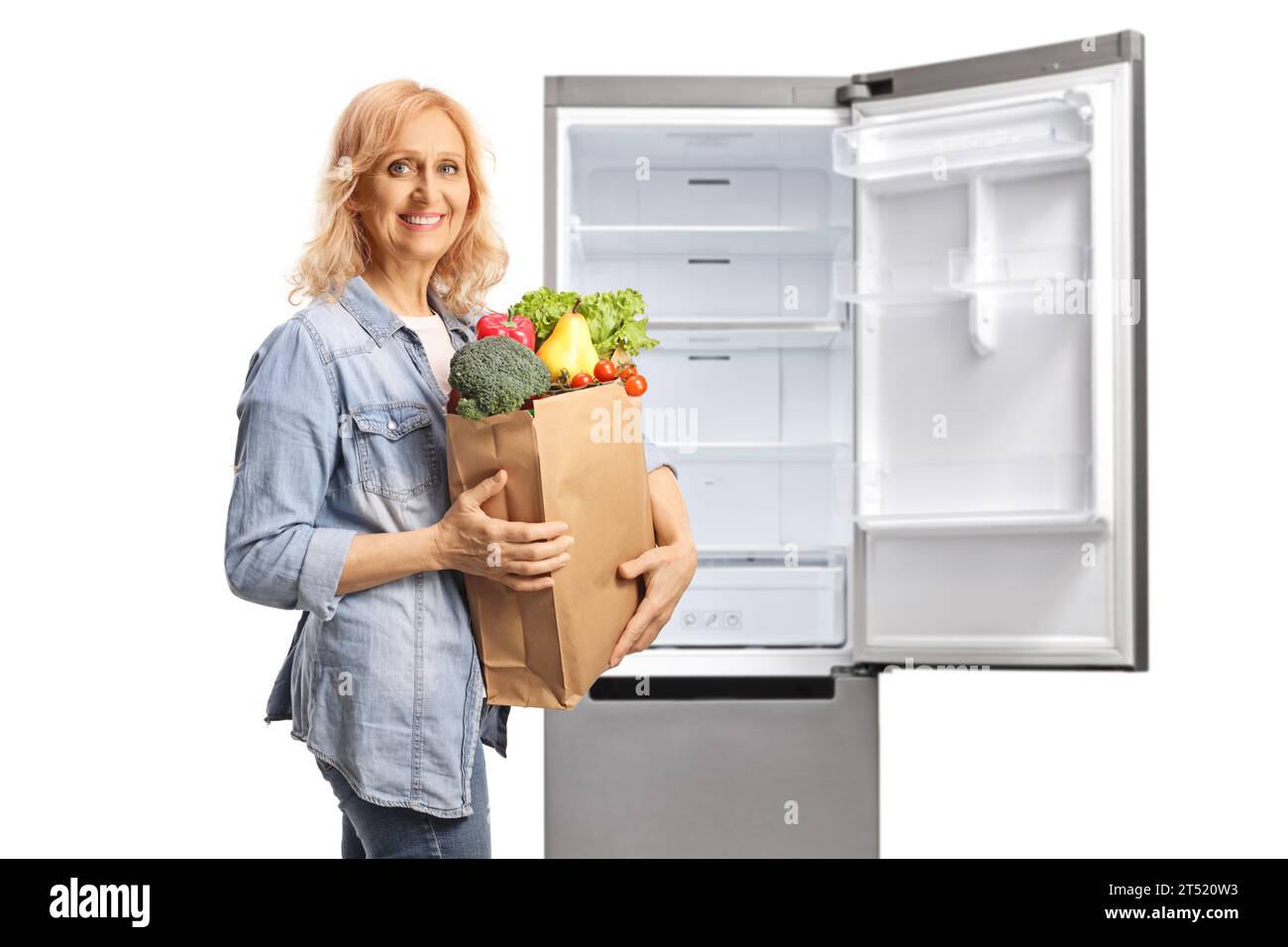 Reife Frau mit einer Lebensmitteltasche neben einem offenen Kühlschrank isoliert auf weißem Hintergrund Stockfoto