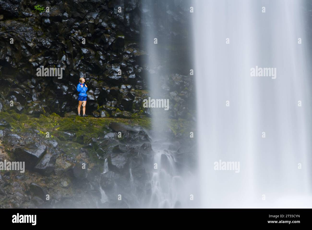 Weibliche Wanderer in einer Regenjacke, die hinter Wasserfällen steht, Hawaii. Stockfoto