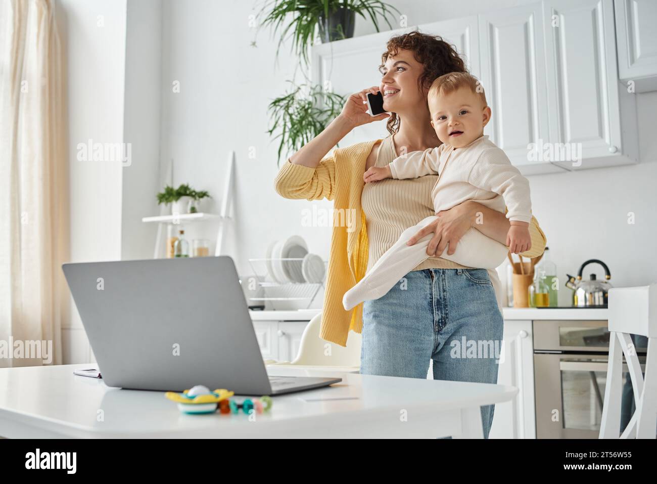 Karriere und Elternschaft, glückliche Frau, die auf dem Smartphone spricht und das Baby in der Nähe des Laptops in der Küche hält Stockfoto