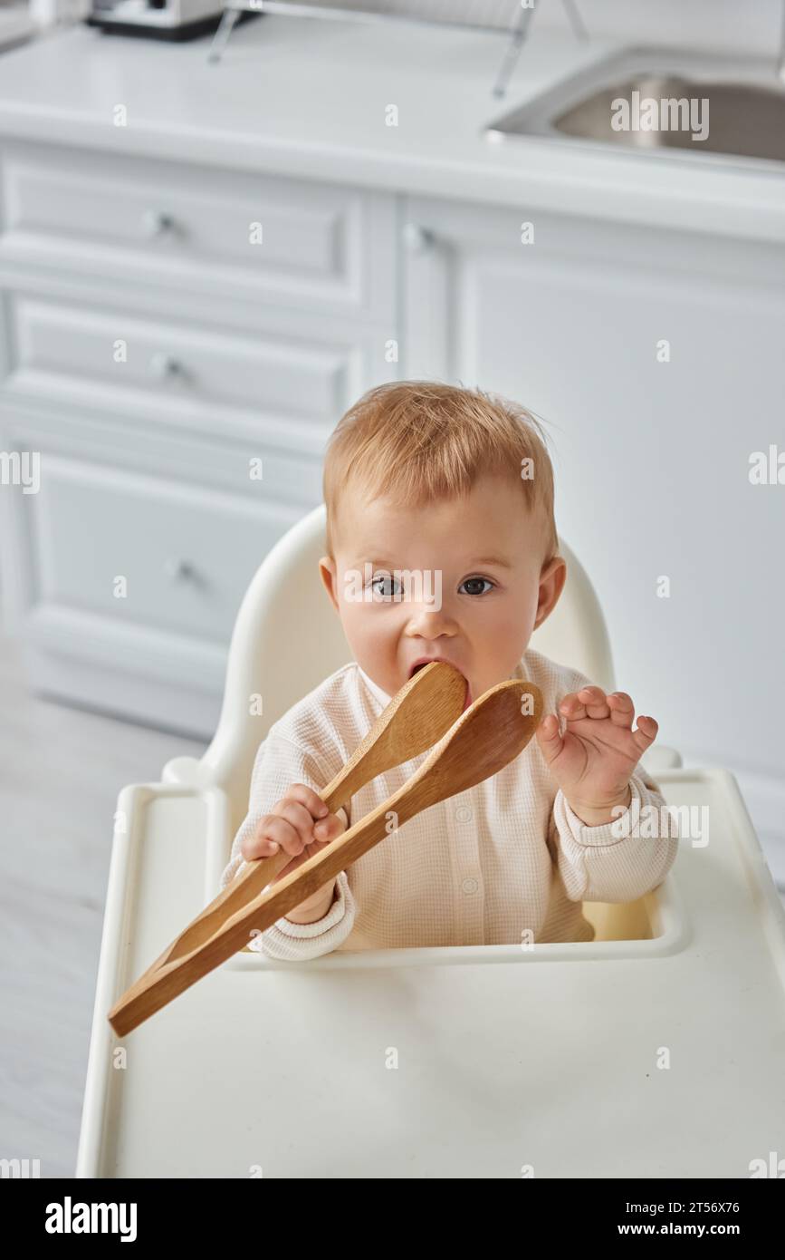 Lustiges Kleinkind Mädchen, das im Kinderstuhl sitzt und hölzerne Kochzange kaut, morgens in der Küche Stockfoto