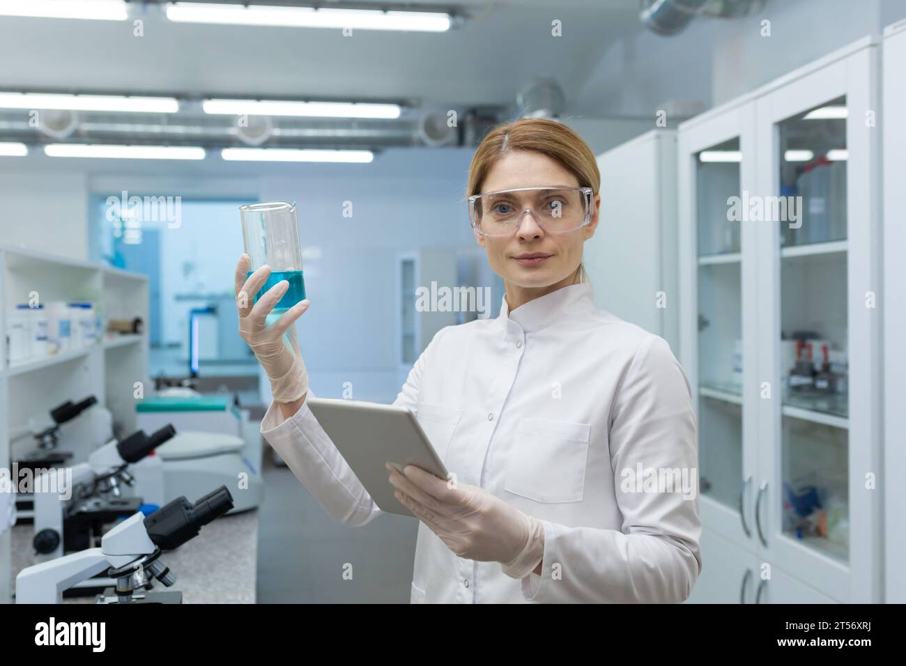 Porträt einer ernsthaften, selbstbewussten Laborarbeiterin, die in die Kamera blickt und mit einem Tablet-Computer in Innenräumen mit Mikroskopen arbeitet. Stockfoto