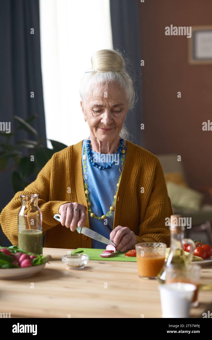 Vertikales Bild einer älteren Frau, die Gemüse für Salat schneidet, während sie am Tisch im Zimmer sitzt Stockfoto