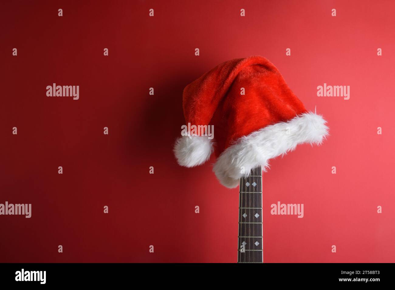 Akustikgitarrenhals mit Weihnachtsmann-Hut auf dem Kopf, der an einer isolierten roten Wand gelehnt ist. Weihnachtsgitarrenmusik-Konzept. Stockfoto