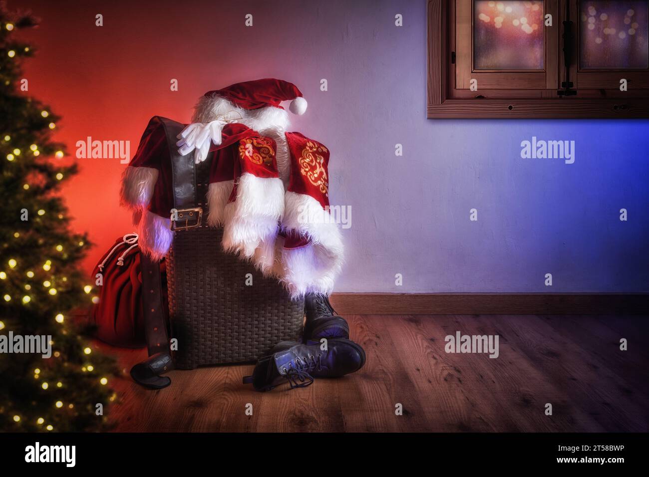 Weihnachtsmann Kleidung und Sack mit Geschenken am Heiligabend in einem hölzernen Raum mit einem Baum mit Lichtern und einem Fenster zum Verteilen. Stockfoto