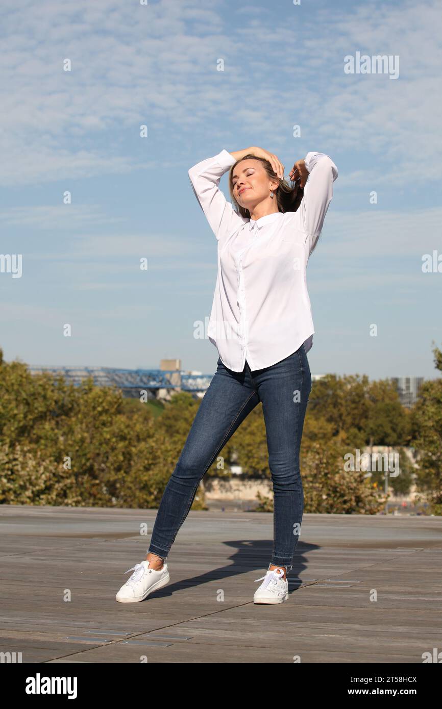 AUSDRÜCKE VON GLÜCK UND WOHLBEFINDEN VON EINEM BLONDEN MÄDCHEN IN WEISSEM HEMD MIT BLAUEM HIMMEL IM HINTERGRUND Stockfoto