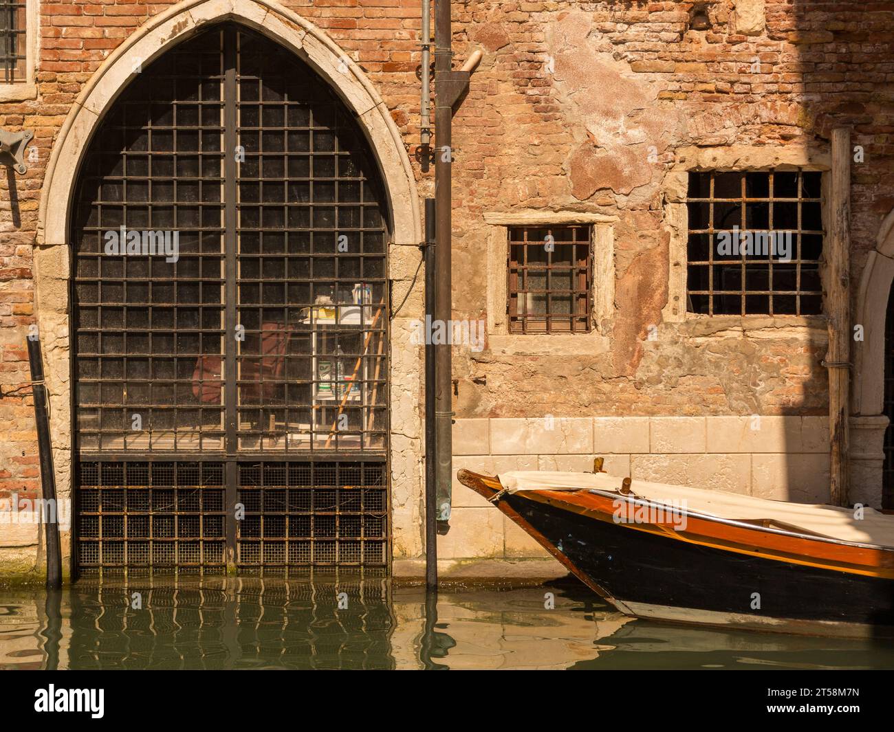Diese ockerfarbene Fassade einer venezianischen Villa ist mit einem Portikus und zwei gegrillten Fenstern dekoriert. Ein mit einer Plane bedecktes Boot ist an seinem verankert Stockfoto