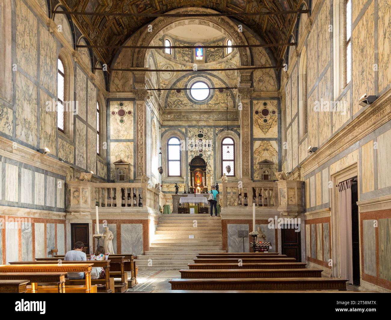 Das Innere einer venezianischen Kirche ist reich mit Marmor dekoriert. Zwei Touristen sitzen in der Kirche, während ein Tourist den Altar betrachtet. Venedig. Stockfoto