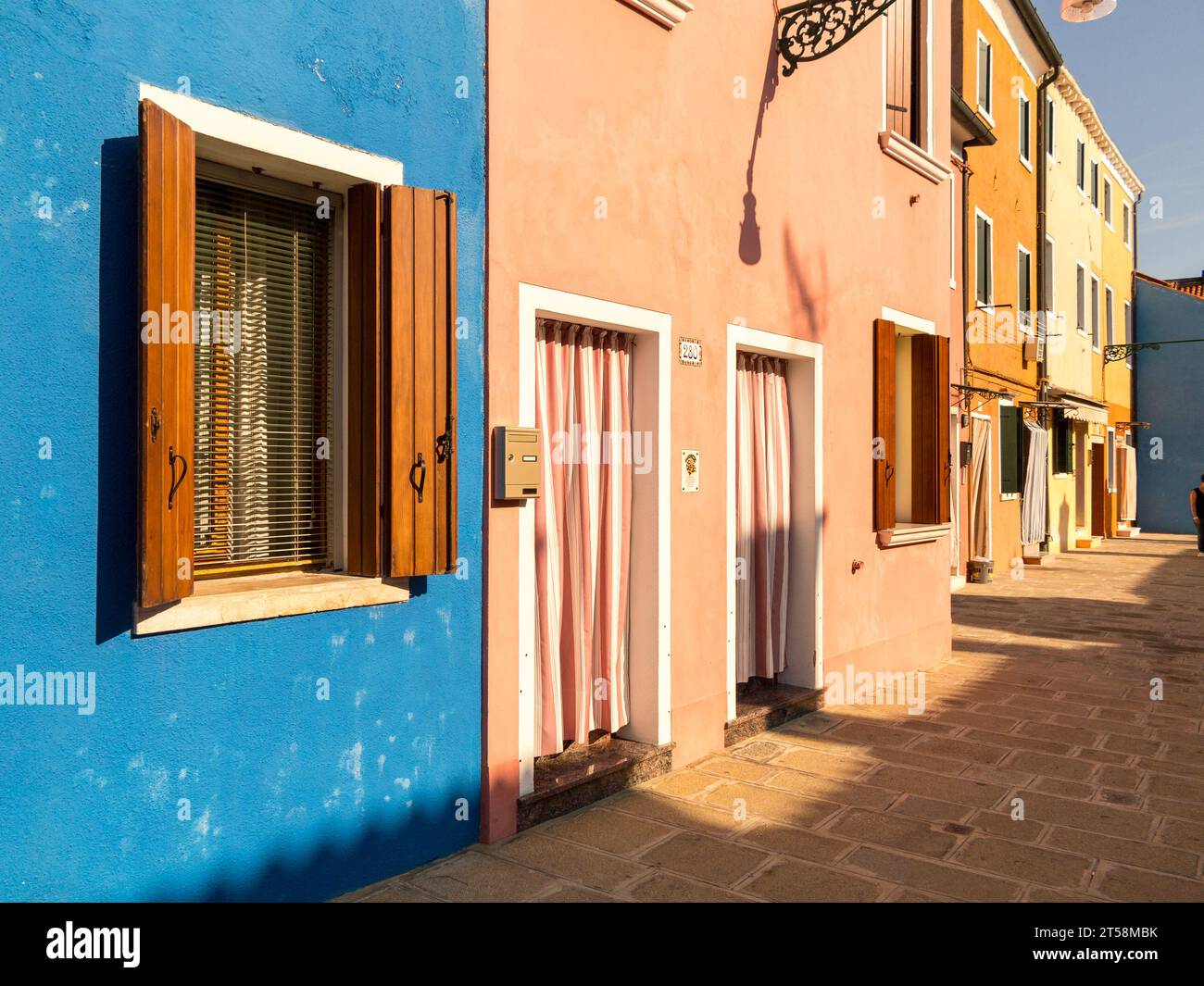 Typische Häuser in Burano, Venedig, Italien. Eine Reihe von blauen, rosa, ockerfarbenen und gelben Häusern überblicken die Straße aus sandfarbenen Kopfsteinpflastern. Stockfoto