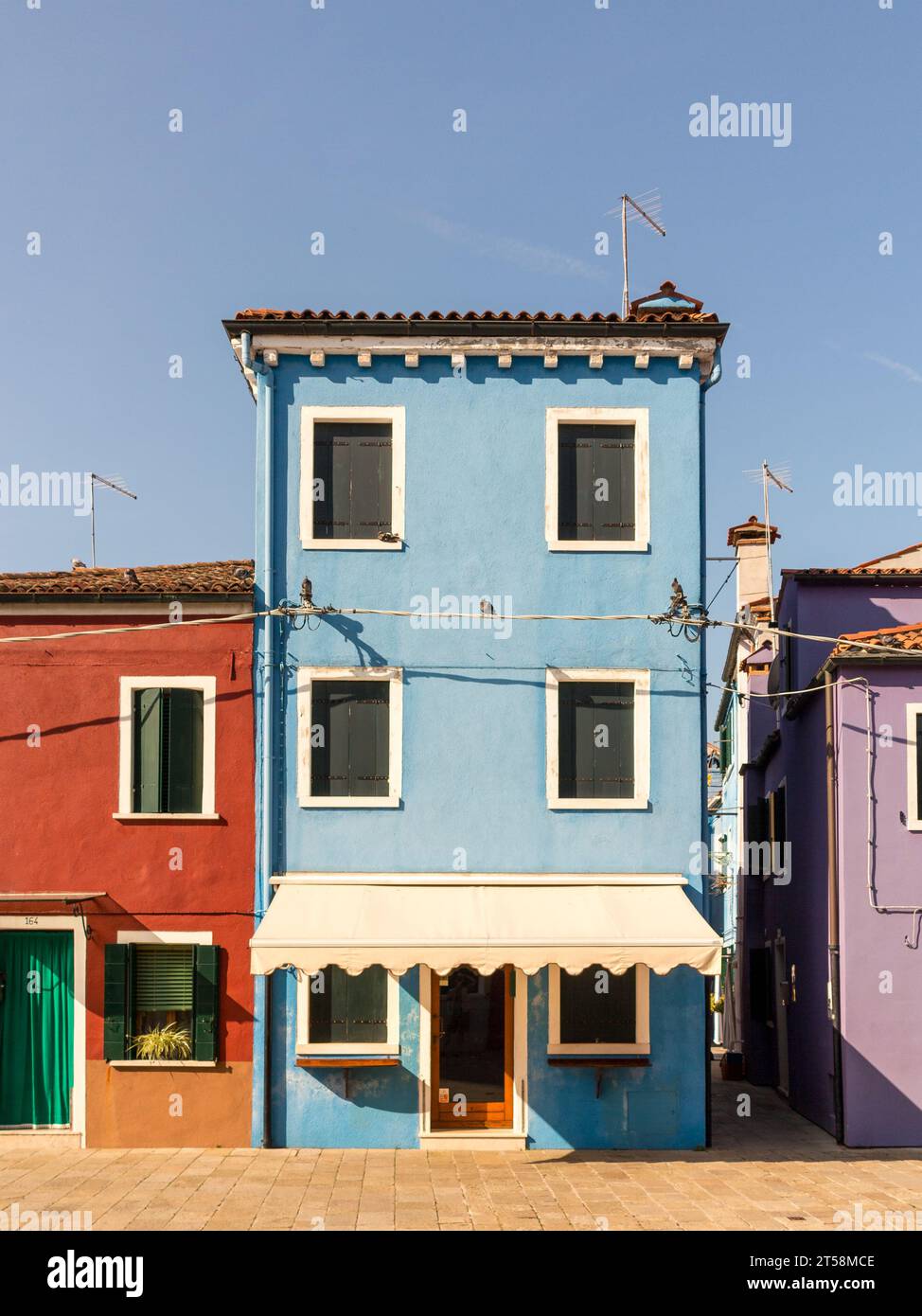 Blaues Haus von Burano in Venedig Italien. Eine Markise ziert die Fassade. Links davon befindet sich ein rostfarbenes Haus und rechts ein lila Haus. Stockfoto