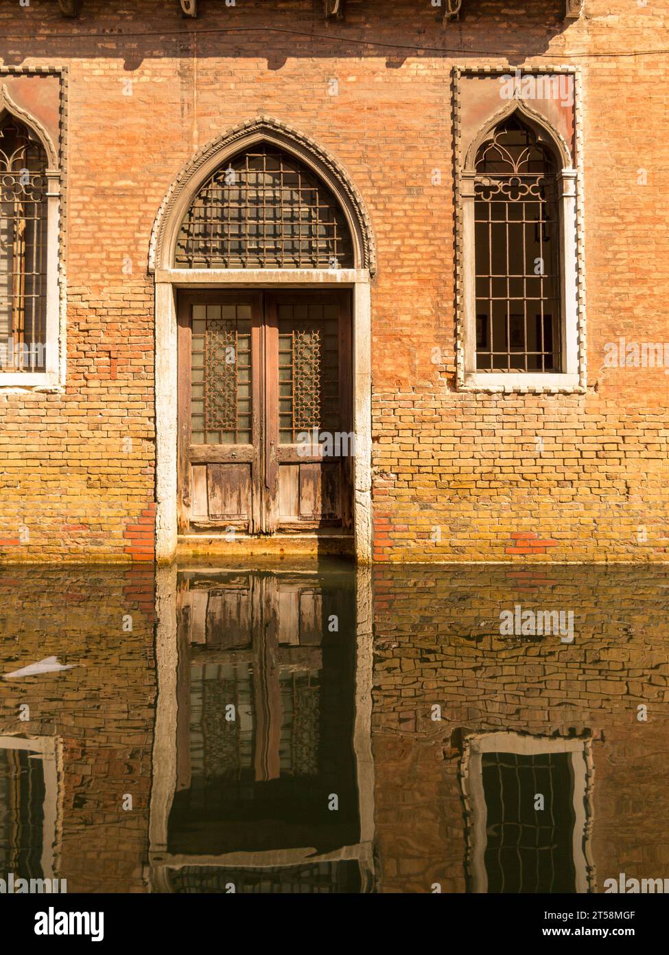 Fassade einer venezianischen Villa spiegelt sich im Wasser des Kanals. Türen und Fenster sind mit dekorativen Grills ausgestattet. Der ockerfarbene Ziegelstein und seine Reflexion c Stockfoto