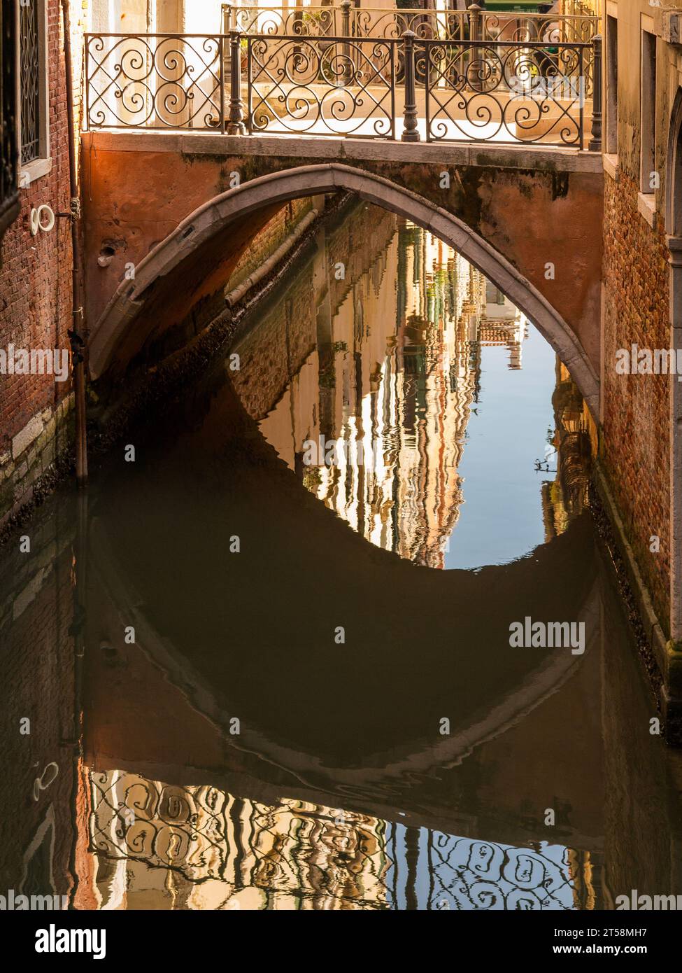 Eine Brücke in Venedig, Italien, spiegelt sich im Wasser eines Kanals. Die Brückenschranken sind mit geometrischen Mustern aus Schmiedeeisen verziert. Die Reflexion Stockfoto